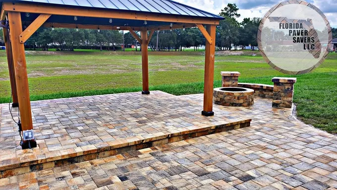 Paver patio beneath a wooden gazebo with a fire pit and seating wall beyond